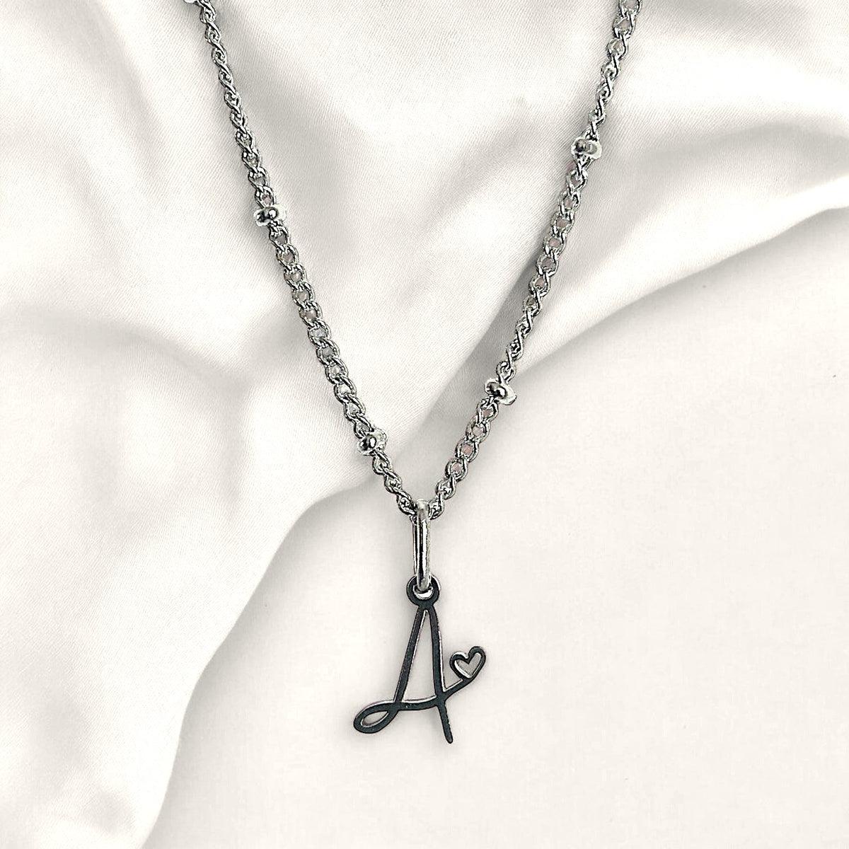 Denoon London cursive initial necklace with heart, silver-tone, letter A shown on cream